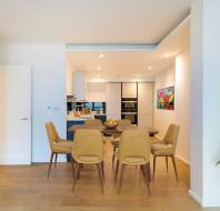 Modern dining area with six mustard-yellow chairs around a wooden table. A kitchen with white cabinets and colorful wall art is visible in the background. A potted plant is on the right.