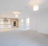 Spacious empty living room with white walls, beige carpet, and an open-plan kitchen featuring light wood cabinets. Two ceiling lights illuminate the space, and there are large windows and a door.