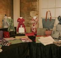 A display of handbags, scarves, and small accessories arranged on black-covered tables in front of a stone wall and a window.