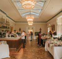 A spacious, elegant room with chandeliers features a bar on the left and several tables displaying goods, with people browsing and sitting throughout the area.
