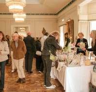 People browse and shop at indoor market tables displaying various goods in a well-lit, elegant room with large windows and chandeliers.