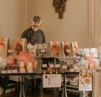 A person stands behind a table displaying various packaged baked goods and sweets for sale, with price tags and signs, in a well-lit indoor setting.