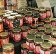 Rows of assorted jam jars with gold lids are displayed on a market table, each labeled with flavors like fine cut orange, rhubarb ginger, and plum & port.