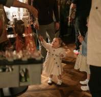 A toddler in a white dress reaches up toward an adult handing out treats in a room with several people and a wooden floor.