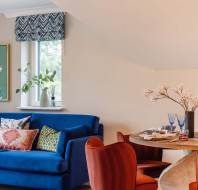 Living room with a blue sofa, colorful cushions, a painting, and a round dining table with red chairs and set tableware. A window with a patterned shade and a vase of flowers is present.