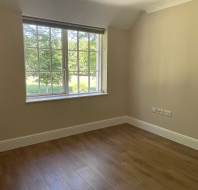 Empty room with beige walls, wooden floor, and a large window letting in natural light; multiple electrical outlets visible on the right wall.