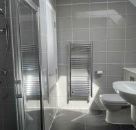 A modern bathroom with gray tiles, a glass shower, a wall-mounted towel radiator, a toilet, and a white sink under natural light.