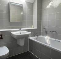 Modern bathroom with gray tiles, wall-mounted toilet, white sink with mirror, bathtub with handheld shower, and a small shelf with a soap dispenser.