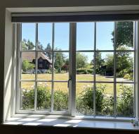 A large window with white frames looks out onto a garden with green grass, blooming flowers, a path, and houses surrounded by trees on a sunny day.
