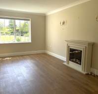 Empty living room with light wooden floors, beige walls, a fireplace, two wall sconces, and a large window letting in natural light.