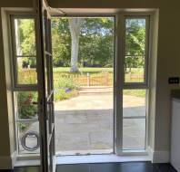 Open glass door leading to a stone patio and garden with trees and a fence visible outside, sunlight illuminating the outdoor area.