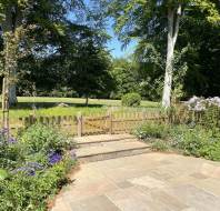 Stone patio with flowering plants borders a wooden picket fence and gate, opening to a grassy field with tall trees under a clear blue sky.