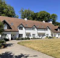 Row of modern, white-painted houses with brown roofs and dormer windows, surrounded by greenery and a neatly maintained lawn under a clear blue sky.