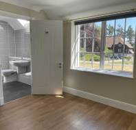 A bright, empty room with wood flooring, a large window, and an open door leading to a modern bathroom with gray tiles and white fixtures.