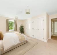 Bright, neutral-toned bedroom with a double bed, bedside tables, two white closets, a window with green curtains, and an open door leading to a hallway.