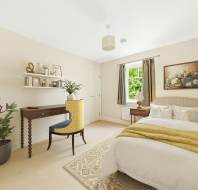 A neatly arranged bedroom with a bed, yellow-patterned chair at a desk, a potted plant, wall shelves with decor, and a window with brown curtains letting in natural light.