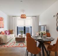 Modern living and dining area with a white sofa, orange chairs, round dining table, patterned rug, and large window with white curtains.