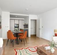 Modern open-plan living and dining area with a white sofa, colorful cushions, orange dining chairs, and a kitchen in the background.