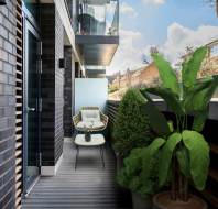 A small modern balcony with a lounge chair, a side table, potted plants, and brick walls, overlooking neighboring buildings and a blue sky.
