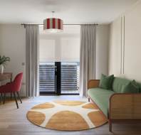A modern living room with a green sofa, round orange and white rug, a wooden desk with a red chair, and glass doors covered by white curtains leading to a small balcony.