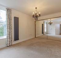 Empty room with beige carpet, white walls, a radiator, chandelier, large mirrored sliding closet doors, and windows with patterned curtains showing greenery outside.