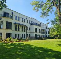 A three-story white apartment building with balconies, surrounded by green grass, trees, and landscaped gardens on a sunny day.