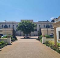 A wide pathway flanked by two white buildings leads to a large central building with balconies, with a leafy tree in the courtyard under a clear blue sky.