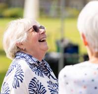 An older woman with short white hair and sunglasses laughs outdoors, facing another person with white hair.