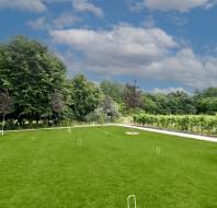 A well-maintained grassy lawn with croquet hoops set up, surrounded by trees and shrubs under a partly cloudy sky. A white bench is visible on the left side.