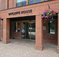 Entrance to Wycliffe House, a red brick building with glass doors, hanging flower baskets, and a paved walkway.