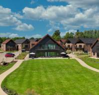 A modern residential complex with several houses, a large central lawn, pathways, parked cars, and a partly cloudy sky.