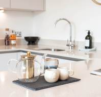 A glass teapot and two ceramic cups on a slate tray sit on a kitchen counter near a sink, with various kitchen items in the background.
