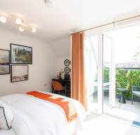A bright bedroom with a bed, desk, and wall art, featuring open glass doors leading to a balcony with greenery and outdoor seating.