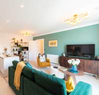 A modern living room with green walls, a green sofa, a yellow armchair, a flat-screen TV, a wooden cabinet, and a round coffee table with flowers.
