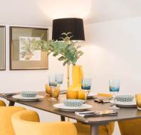 A dining table set for six with blue and gold glassware, patterned bowls, yellow chairs, a yellow vase with greenery, and framed art on the wall.