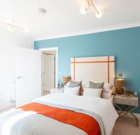 Modern bedroom with a blue accent wall, upholstered headboard, two bedside tables with lamps, and a neatly made bed with white and orange bedding.