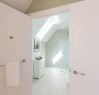 A modern white bathroom with a skylight, tiled floor, glass shower, small cabinet, wall art, and a towel hanging on the door.