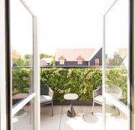 Small balcony with two black chairs and a round table, facing a green privacy wall, with rooftops and trees visible in the background.