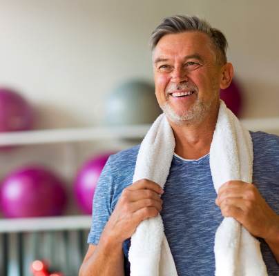 Smiling older man in a blue shirt holds a white towel around his neck in a gym, with colorful exercise balls in the background.
