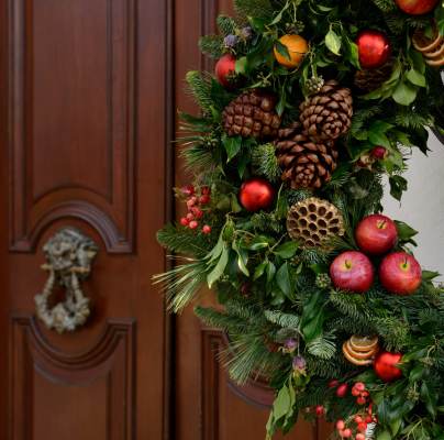 A festive wreath made of pinecones, apples, berries, and greenery hangs on a wall next to a wooden door with a decorative metal knocker.