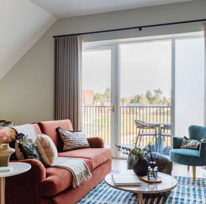 Modern living room with a red sofa, teal armchair, and coffee table on a patterned rug. Large windows open to a balcony with a table and chairs. Dining area visible in the background.