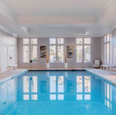 Indoor swimming pool with clear blue water, surrounded by beige tiles. Large windows let in natural light. Several lounge chairs and tables are placed along the sides.
