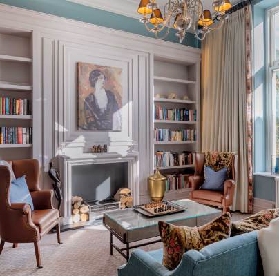 A cozy living room with bookshelves, a fireplace, and armchairs, featuring a large window and a chandelier.