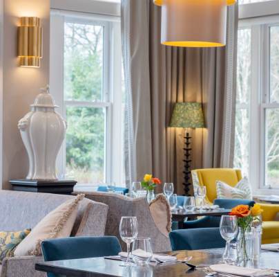 A modern dining room with chairs, tables set with glassware, and bright natural light from large windows. Vases and lamps decorate the space.