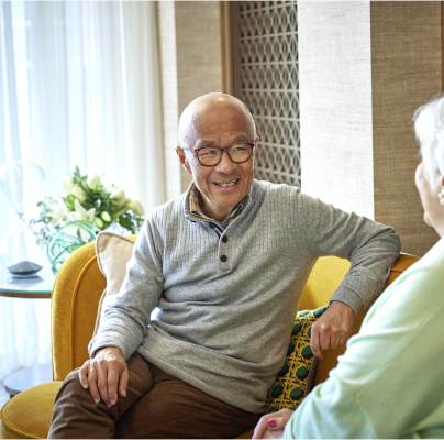 Two elderly people sitting on a yellow sofa, engaged in conversation, with light shining through sheer curtains in the background.