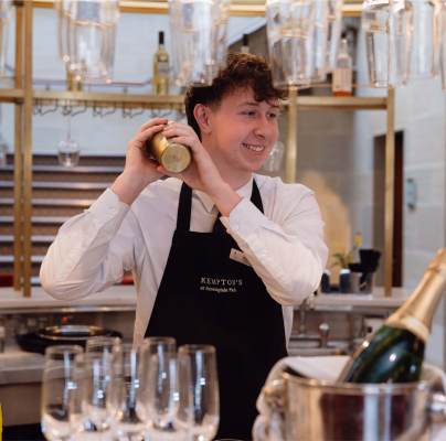 Bartender in a black apron smiles while shaking a cocktail mixer behind a bar. Hanging glasses and a champagne bottle are in the foreground.