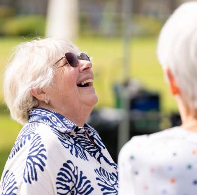 An older woman with short white hair and sunglasses laughs outdoors, facing another person with white hair.
