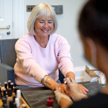 An older woman with gray hair and a pink sweater smiles while receiving a manicure at a nail salon. Bottles of nail polish are visible on the table.