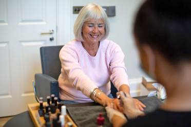 An older woman sits at a table smiling while receiving a manicure from a nail technician in a salon setting.
