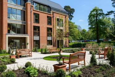 A modern brick apartment building with balconies overlooks a landscaped courtyard featuring benches, young trees, and various potted plants on a sunny day.